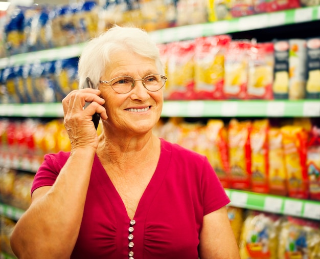 A smiling senior woman holding a discount card while shopping at a grocery store, looking pleased. The background is a blur of shelves and other shoppers.