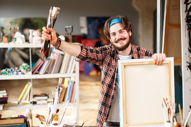 A person smiling while holding a freshly painted wooden frame. Various paint supplies and drop cloths are visible in the background, suggesting a recently completed DIY project.